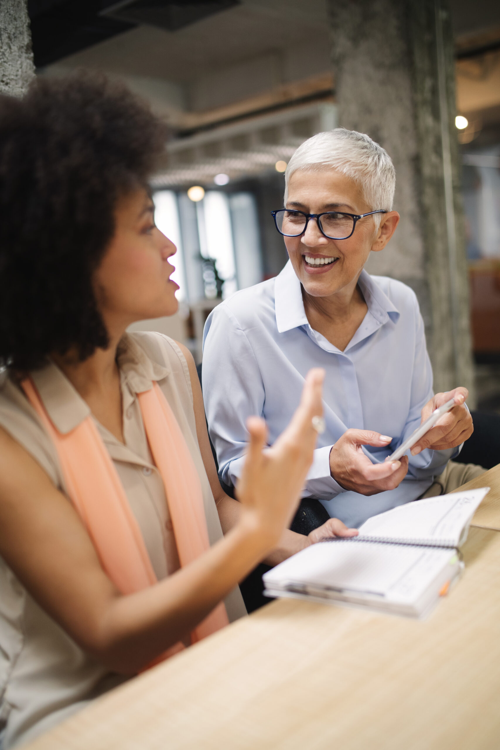 Older businesswoman with short gray hair and glasses smiling while listening to a younger colleague during a professional mentoring conversation in a modern office.